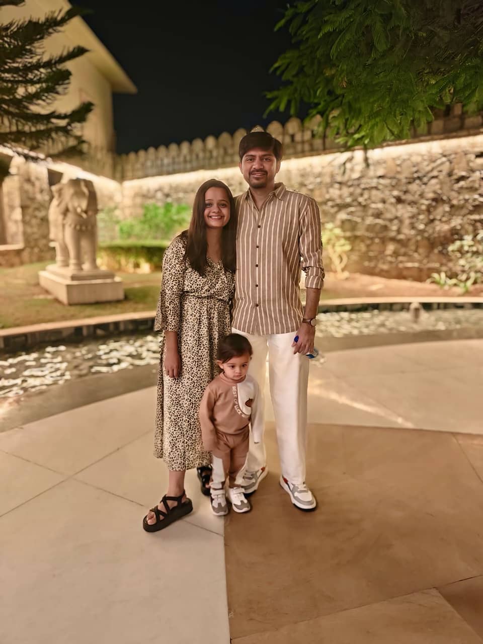 Sagar Shah, on the right, standing outdoors in the evening with his family in front of a floodlit stone wall, wearing a brown-striped shirt and white trousers.